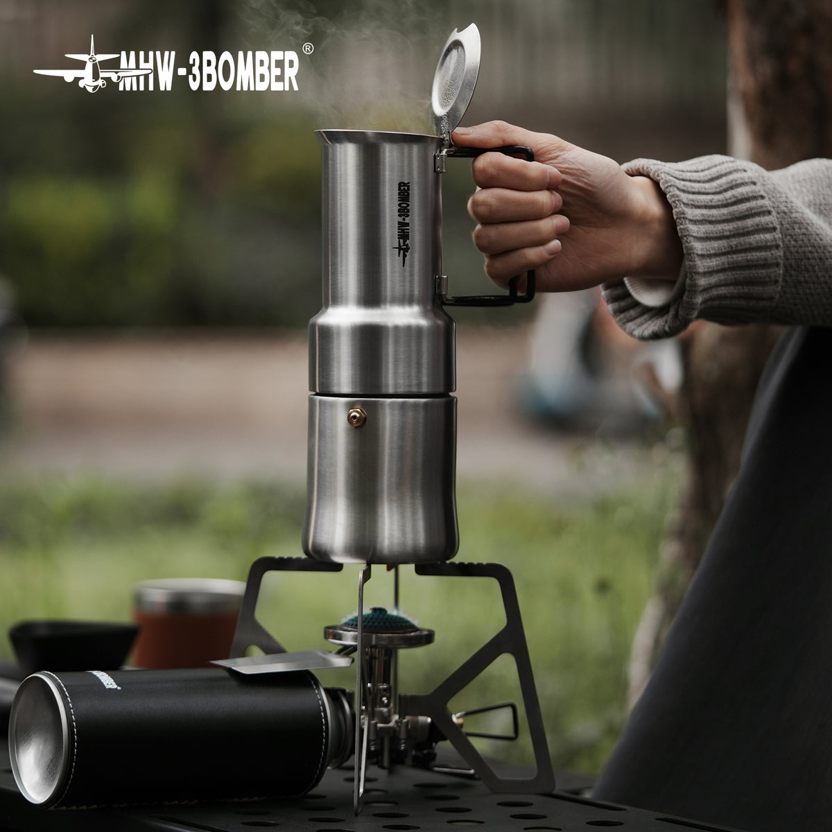MHW-3BOMBER CS6049 MET stove in a separate outdoor coffee-station photograph, showing supports and body beneath a reference pot; pot, cup, fuel cartridge and furniture are not included