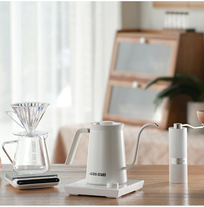 White Assassin electric gooseneck kettle on its matching base beside coffee brewing tools