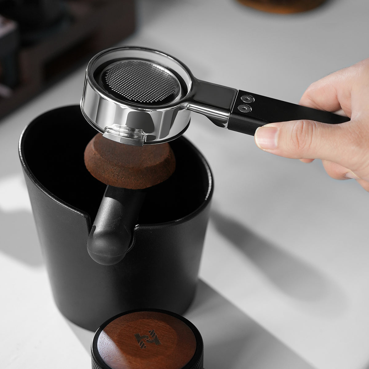 HiBREW H21 Magician 58mm Portafilter — puck removal above a knock box. Scene props are not included unless quoted.