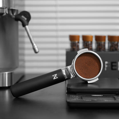 HiBREW H21 Magician 58mm Portafilter — hand-held basket view beside an espresso machine. Scene props are not included unless quoted.
