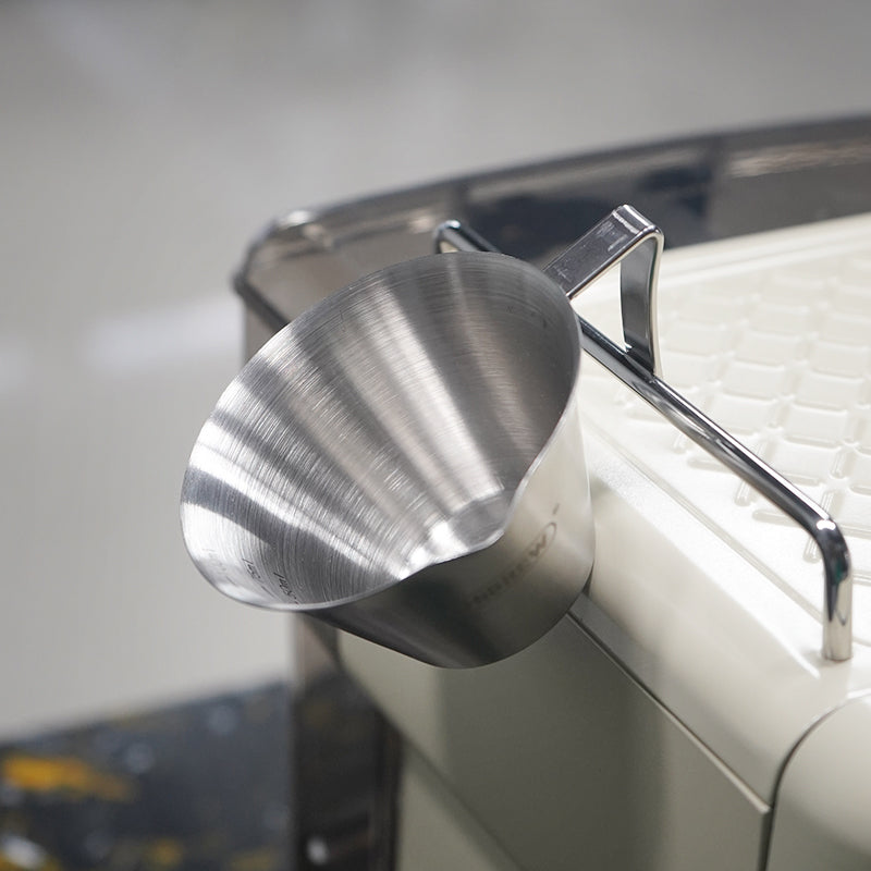 HiBREW 100ml Espresso Measuring Pitcher — tilted view beside an espresso machine. Demonstration props are not included.
