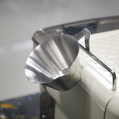 HiBREW 100ml Espresso Measuring Pitcher — tilted view beside an espresso machine. Demonstration props are not included.
