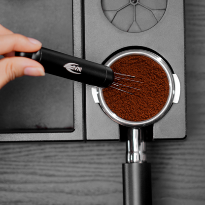 IKAPE black retractable WDT tool distributing espresso grounds, overhead view; portafilter and mat shown separately