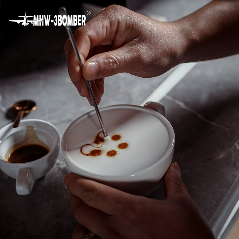 MHW-3BOMBER C5080W Beige White 280 ml latte cup held during a latte-art demonstration; art tool and other cups are photo props.
