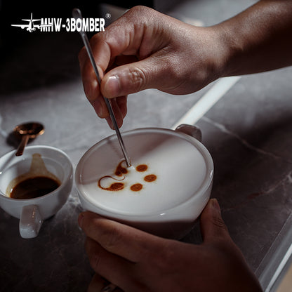 MHW-3BOMBER C5080W Beige White 280 ml latte cup held during a latte-art demonstration; art tool and other cups are photo props.