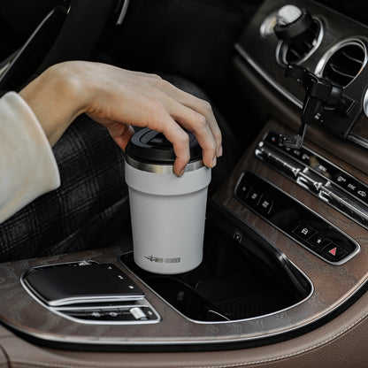 MHW-3BOMBER M5716W Cooki 360 ml travel cup: beige-white cup held in a car setting; other pictured equipment and props are not included.