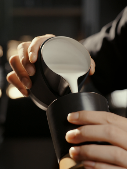 MHW-3BOMBER P6513B black 400 ml GT Handleless Milk Pitcher pouring latte art into a black cup; cup is not included