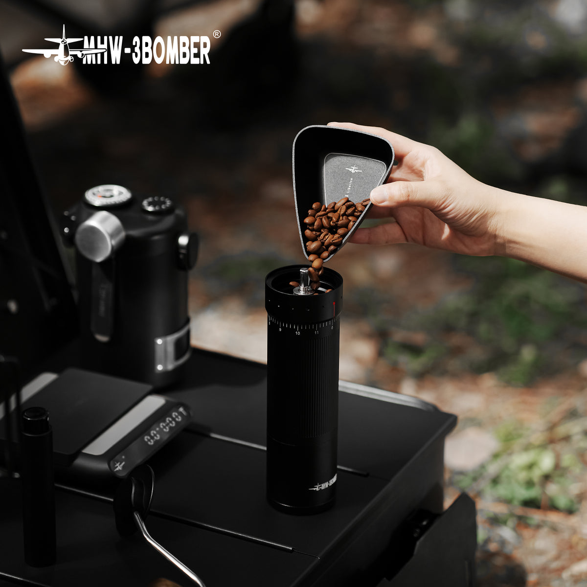 MHW-3BOMBER P6562B small black BeanBoat tray pouring beans into a hand grinder outdoors; grinder and coffee are not included