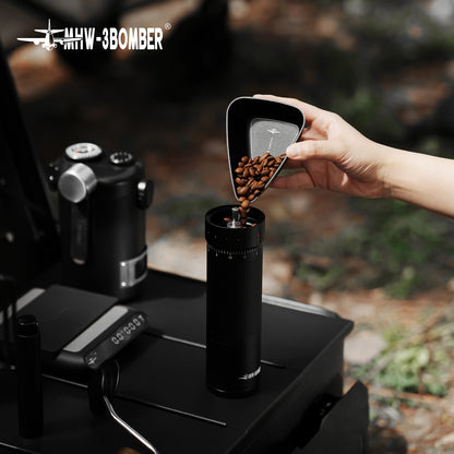 MHW-3BOMBER P6562B small black BeanBoat tray pouring beans into a hand grinder outdoors; grinder and coffee are not included