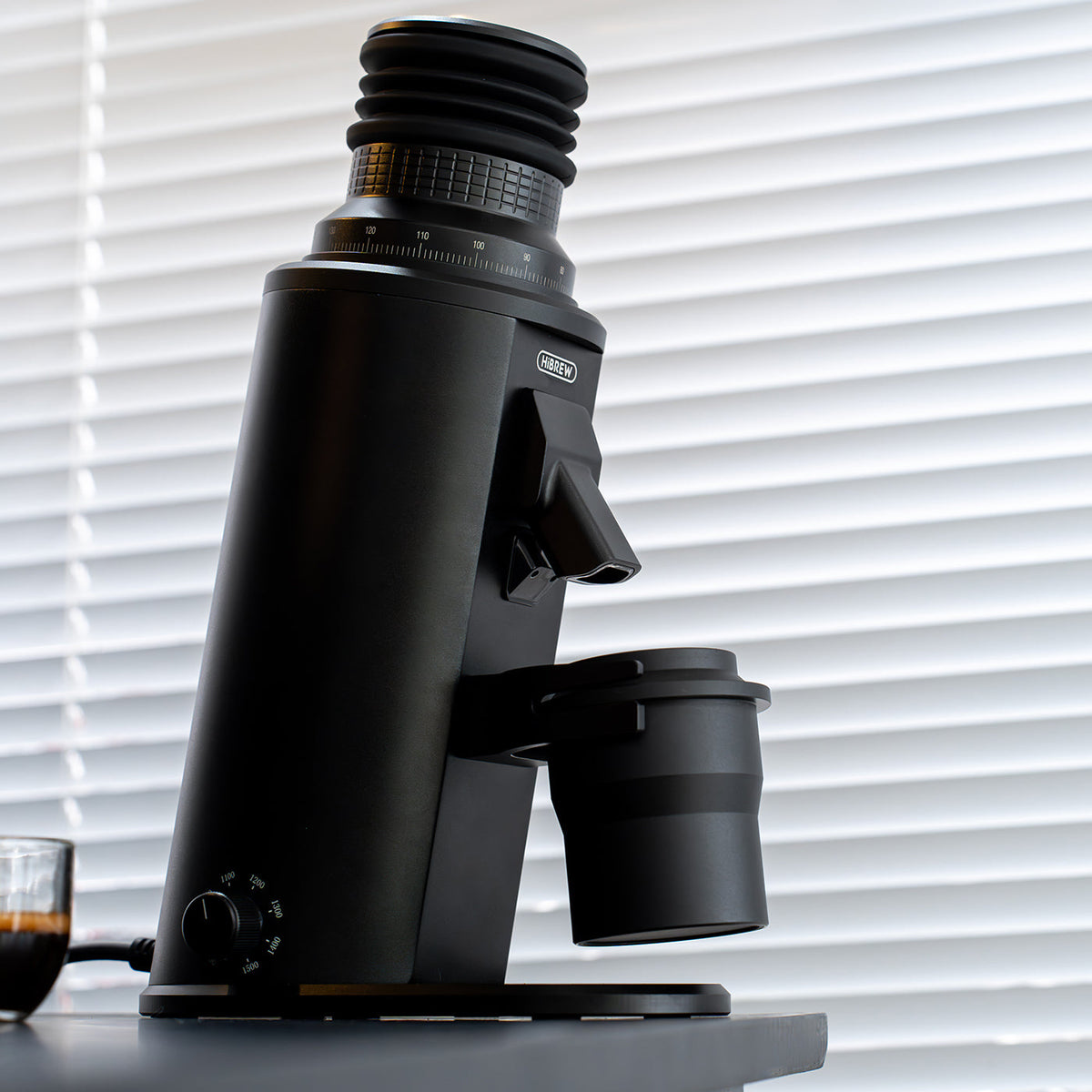 HiBREW G64 Plus black coffee grinder with dosing cup beside a window; glass and coffee are scene props