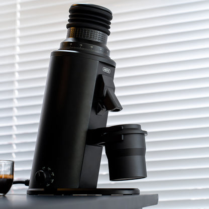 HiBREW G64 Plus black coffee grinder with dosing cup beside a window; glass and coffee are scene props