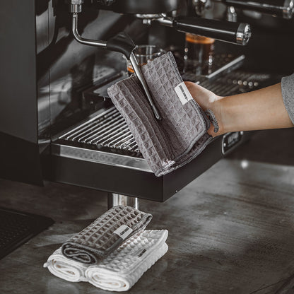 MHW-3BOMBER dark-grey square waffle towel with hanging loop shown wiping a steam wand; background towels are props.