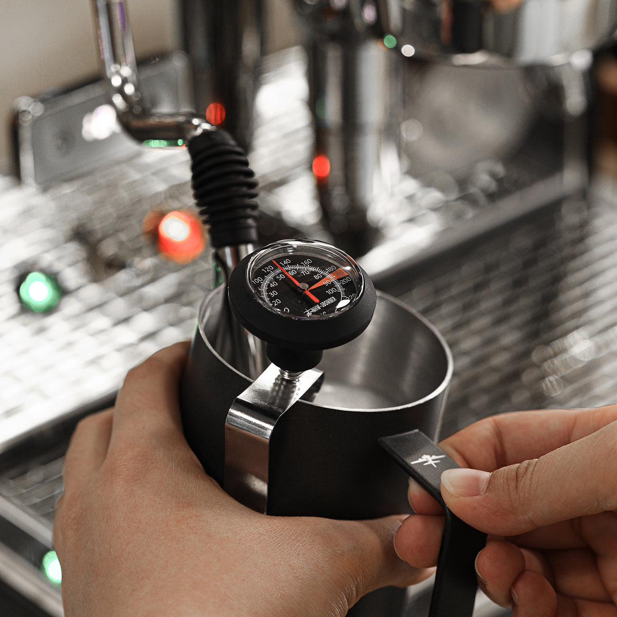 MHW-3BOMBER CT5615B black dial thermometer clipped to a milk pitcher during steaming; pitcher and coffee machine are scene props
