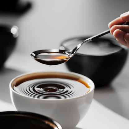 MHW-3BOMBER S5706S black-handle silver-bowl cupping spoon held over a coffee cup; other items are scene props