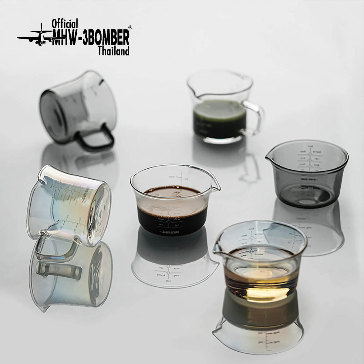 MHW-3BOMBER measuring-cup comparison: clear G5052 single-spout with coffee at centre and clear G5059 handled double-spout at rear; one selected cup supplied