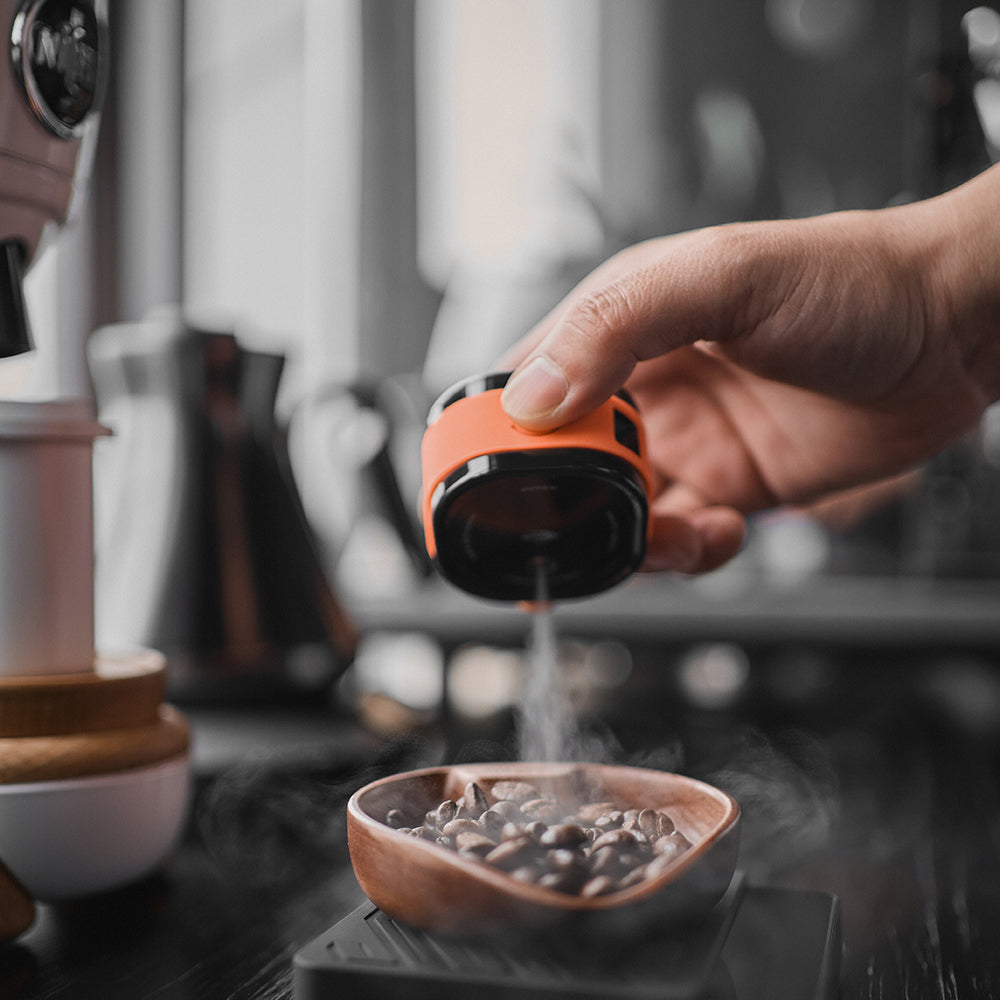 IKAPE electric coffee bean atomizer, black and orange, spraying beans beside grinder