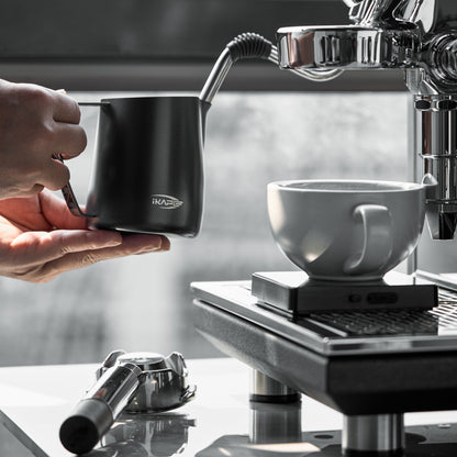Black IKAPE hollow-handle milk pitcher held beneath an espresso-machine steam wand
