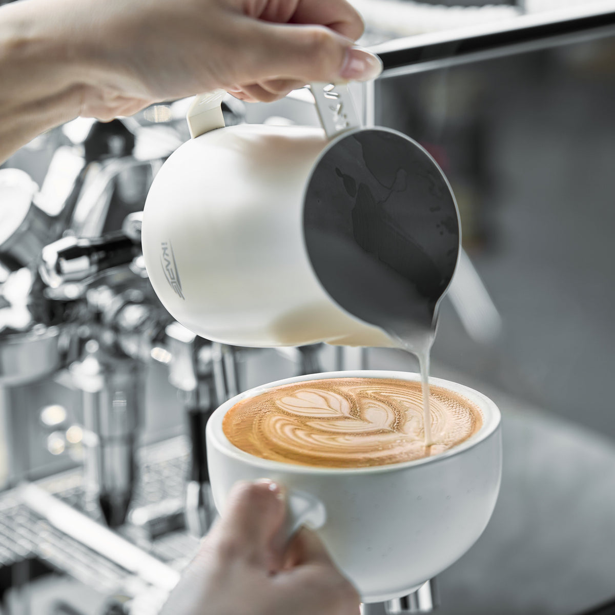 White IKAPE hollow-handle milk pitcher pouring latte art beside an espresso machine