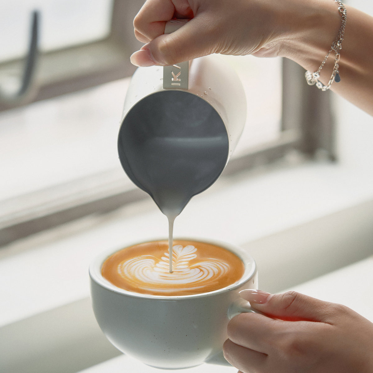 White IKAPE hollow-handle milk pitcher pouring latte art by a window