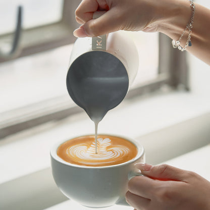 White IKAPE hollow-handle milk pitcher pouring latte art by a window