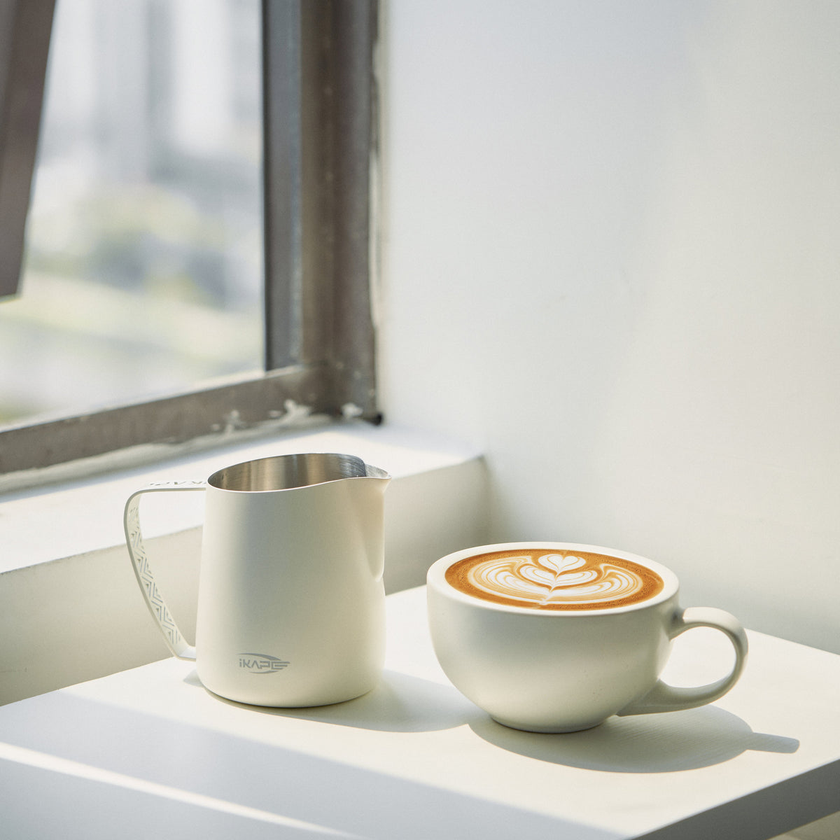 White IKAPE hollow-handle milk pitcher beside a finished latte