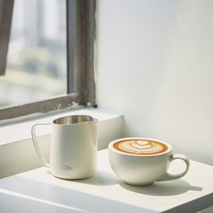 White IKAPE hollow-handle milk pitcher beside a finished latte