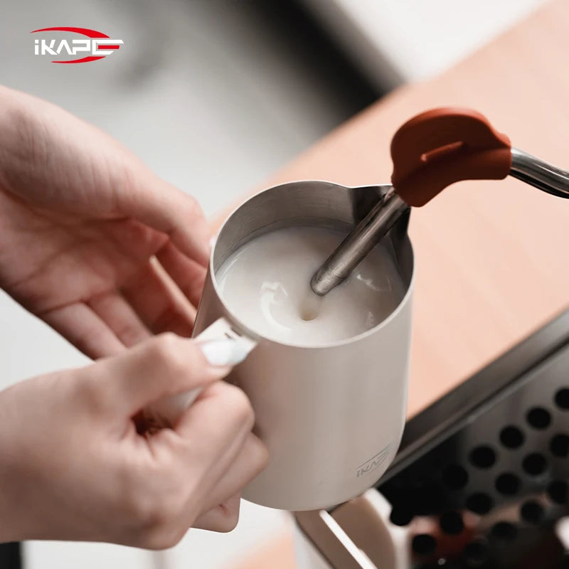White IKAPE hollow-handle milk pitcher steaming milk beside an espresso machine
