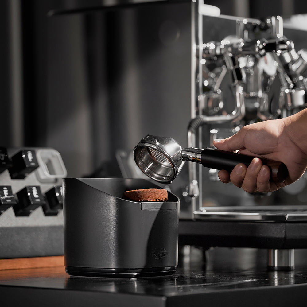 IKAPE V2 black knock box beside an espresso machine during puck removal; machine excluded