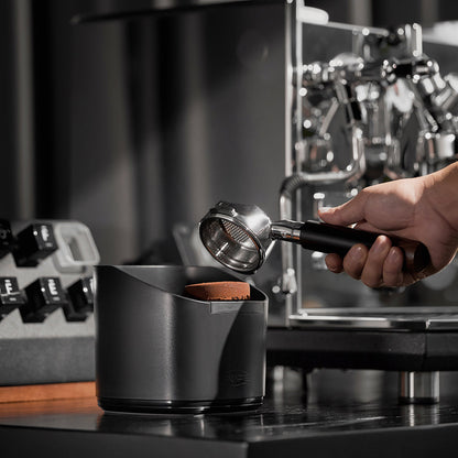 IKAPE V2 black knock box beside an espresso machine during puck removal; machine excluded