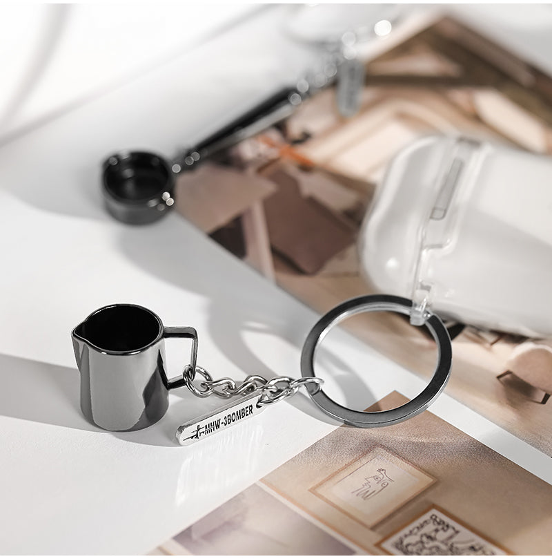 MHW-3BOMBER KC5700 miniature milk-pitcher keychain resting on a book; coffee tools are props