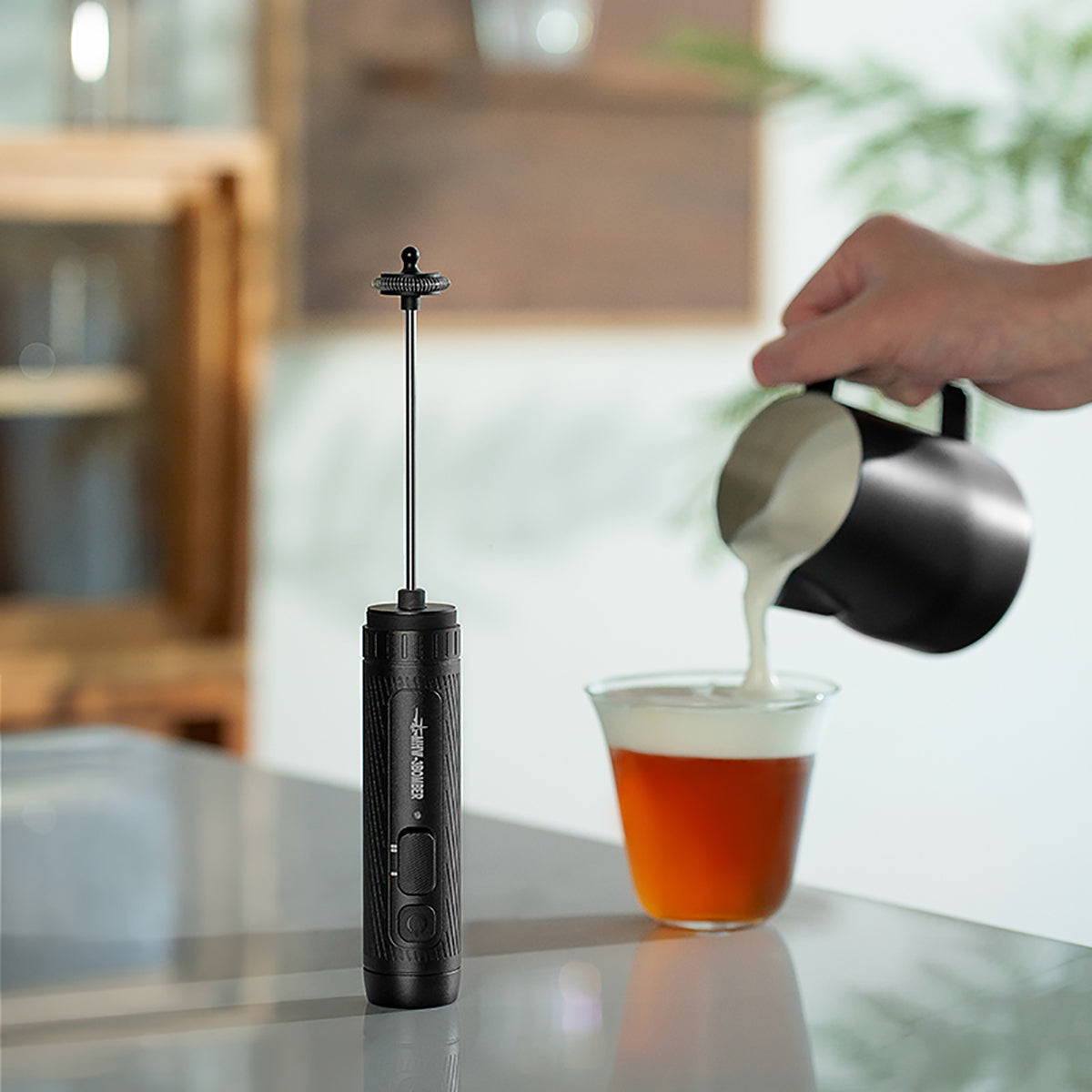 MHW-3BOMBER MF5893 black Magician frother standing beside a drink while milk is poured; pitcher and drink not included