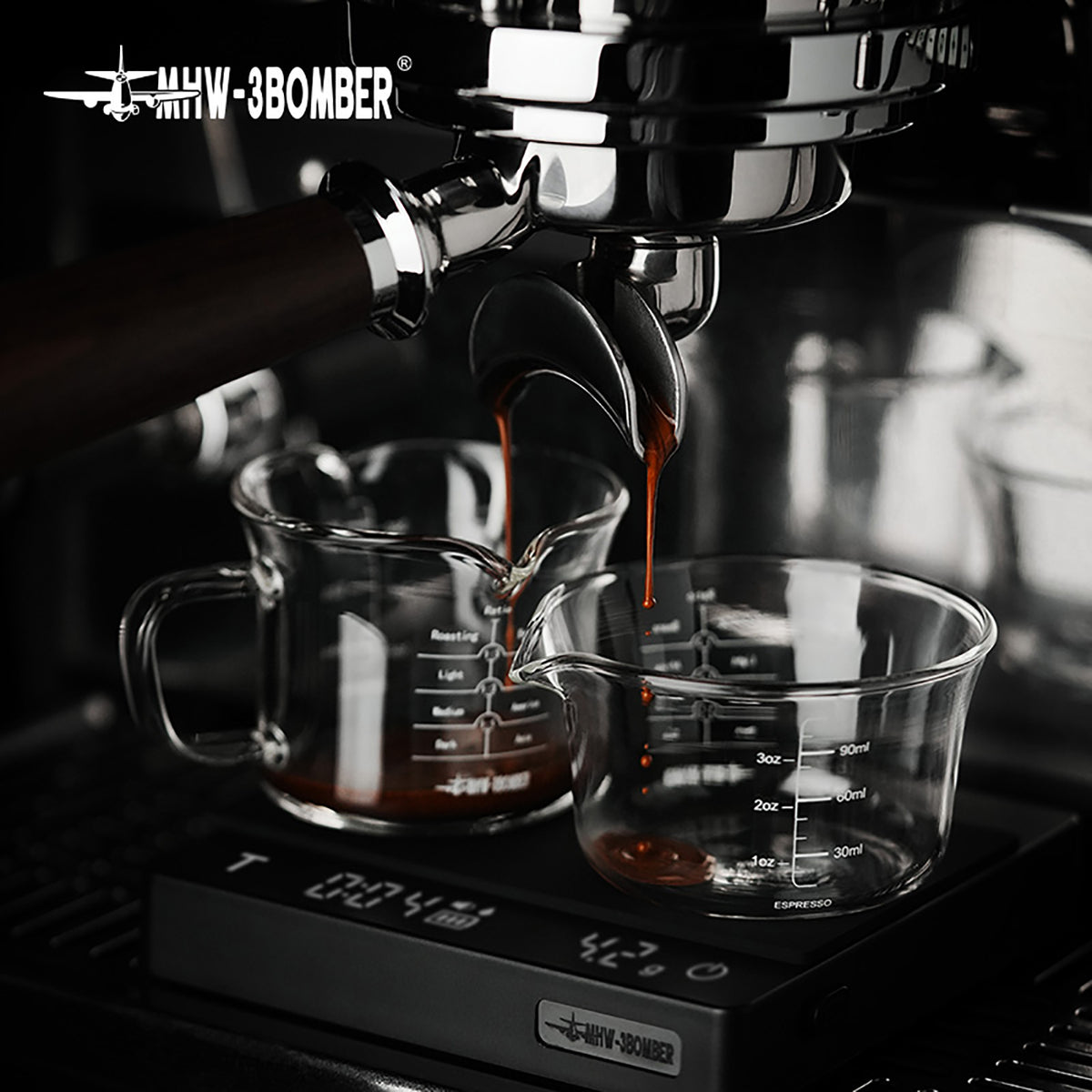 Clear G5059 handled double-spout cup on the left and G5052 single-spout cup on the right in a shared espresso scene; configurations are sold separately, machine and scale are props