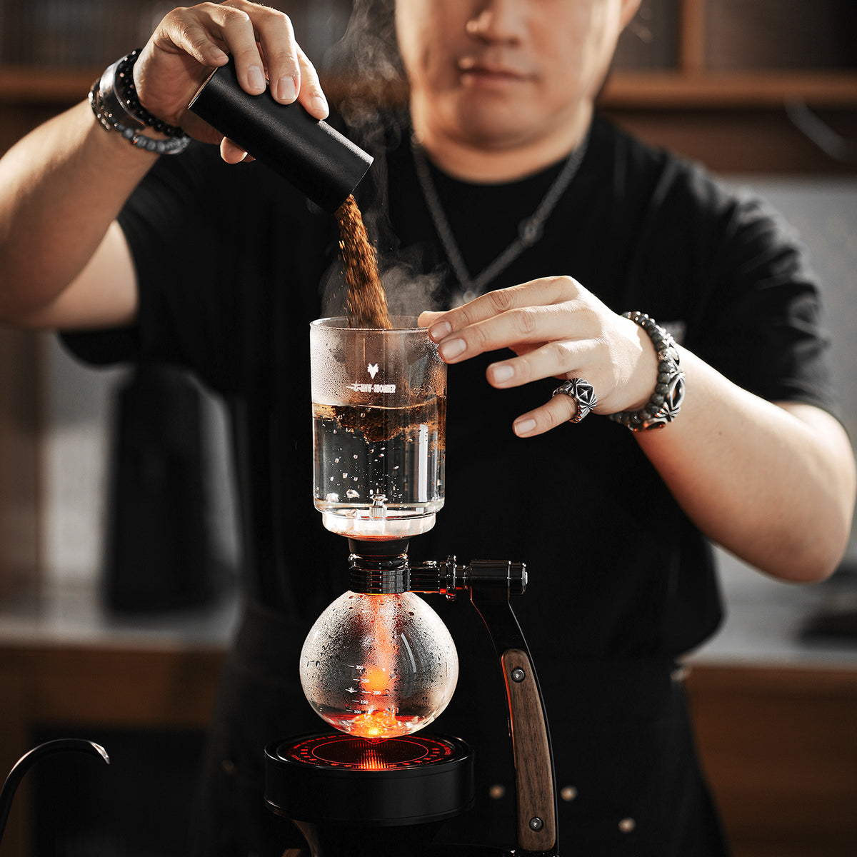 MHW-3BOMBER S5916 Twilight siphon brewer with ground coffee being added to the upper glass chamber; heating equipment and other scene props are not included