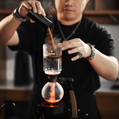 MHW-3BOMBER S5916 Twilight siphon brewer with ground coffee being added to the upper glass chamber; heating equipment and other scene props are not included