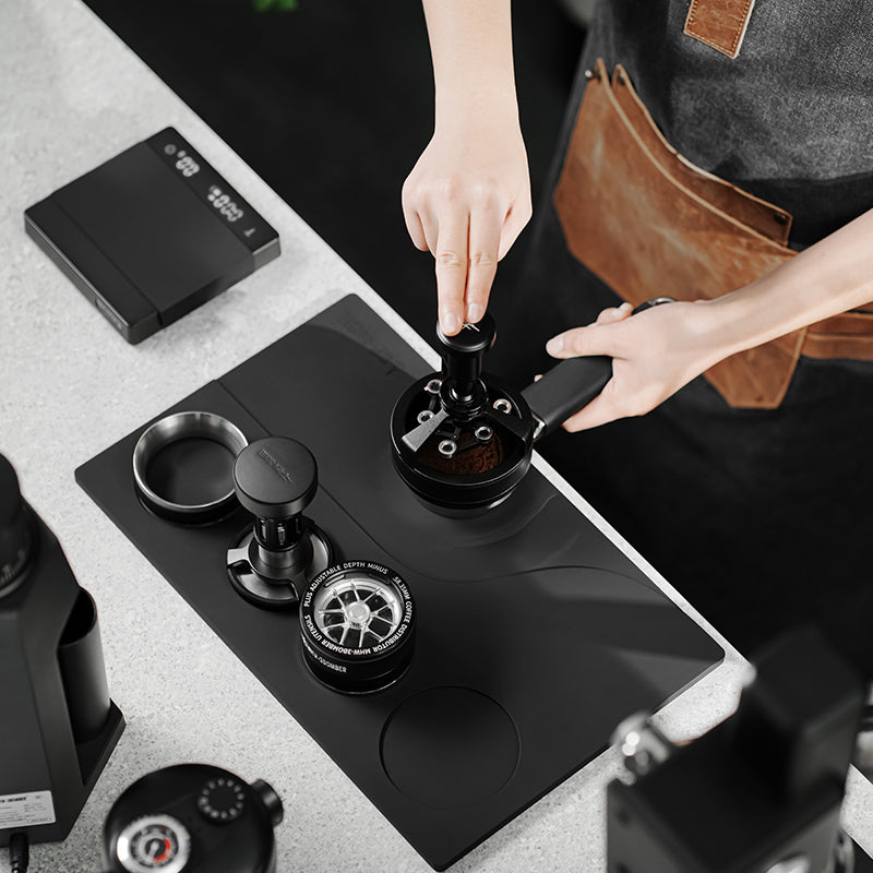 MHW-3BOMBER SP5538 Black recessed Yu tamping mat during coffee distribution at a barista workstation; tools and appliances are scene props, not included.