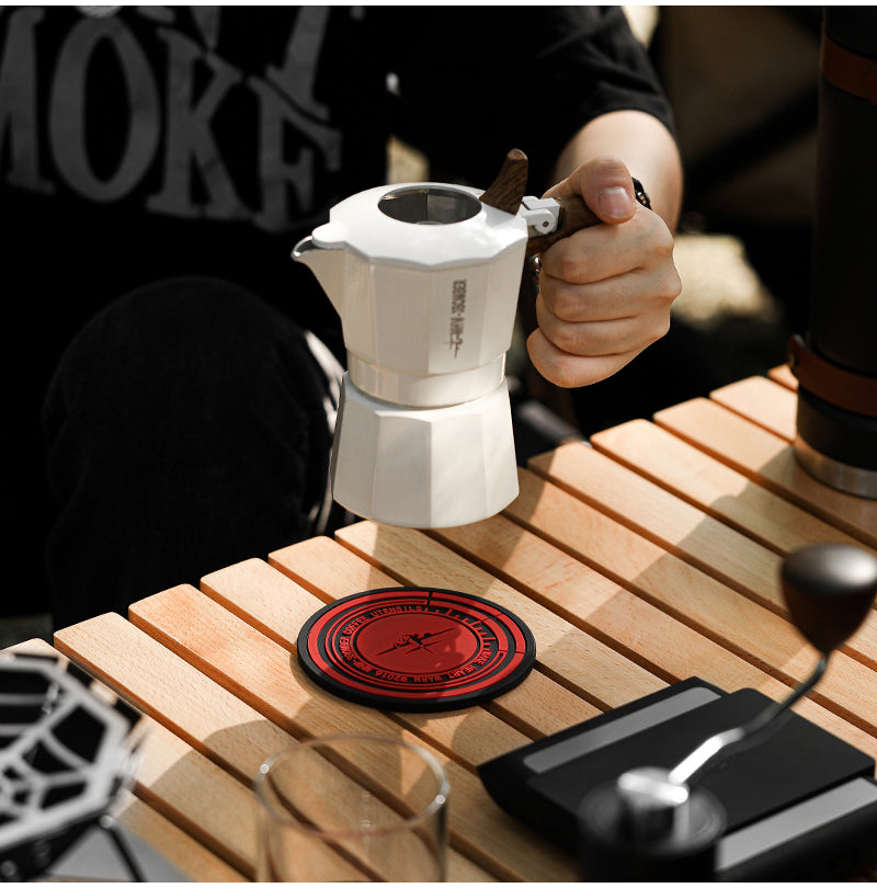 MHW-3BOMBER moka pot being lifted from a mat on a wood counter; pot separate