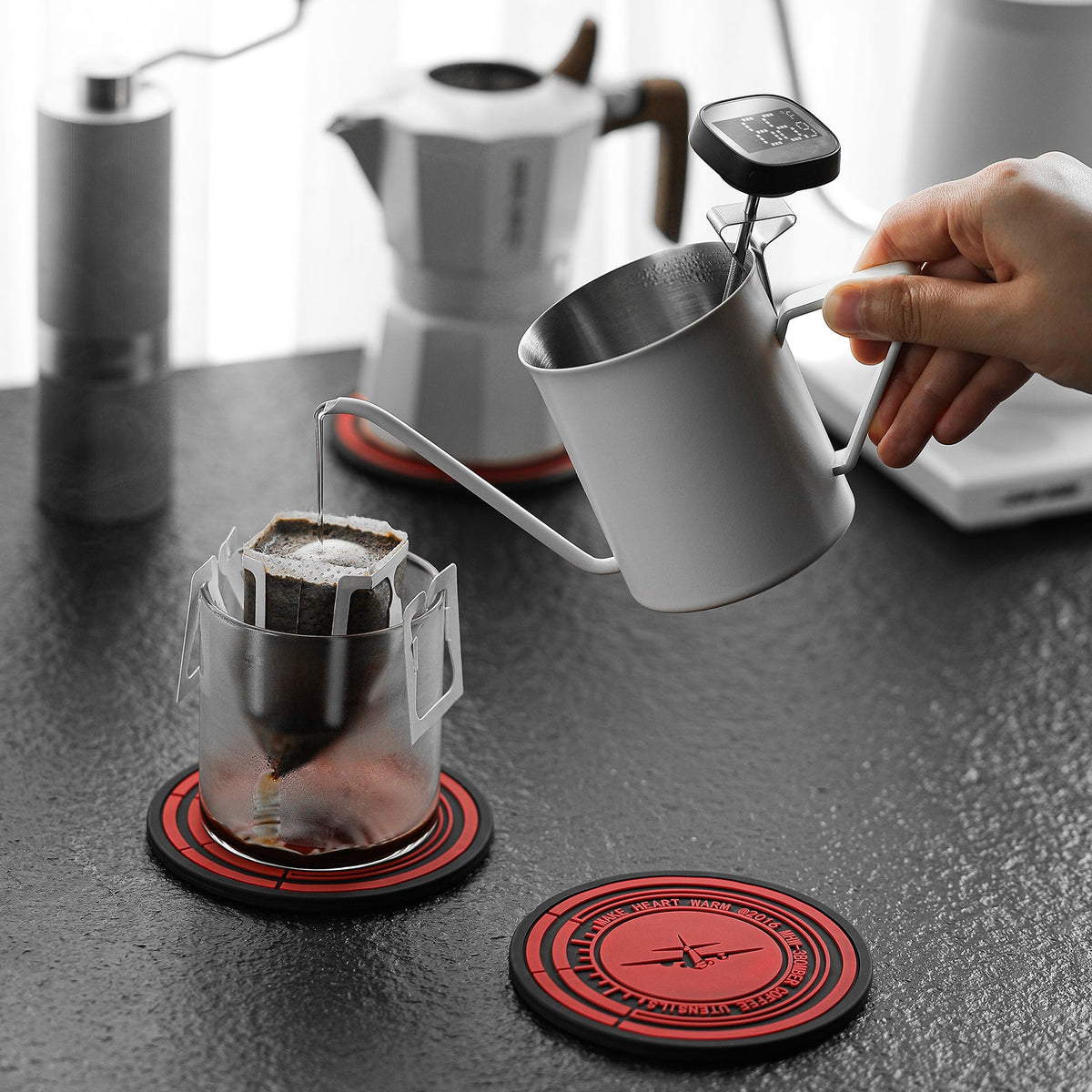 MHW-3BOMBER SP5885 red-black silicone mat under a coffee dripper during pouring; dripper, kettle, cups and additional mat are props